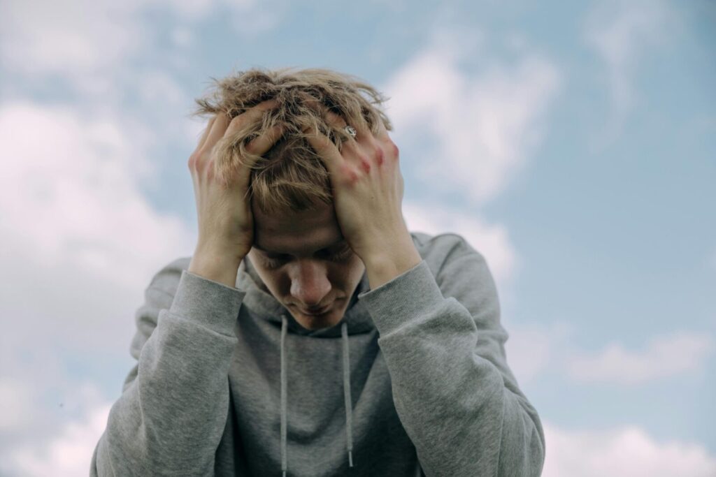 Young man in gray hoodie holds head in frustration, set against cloudy sky.
